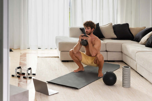 Man performing kettlebell squat in home gym setup with adjustable weights, exercise mat, and laptop for home workout convenience.