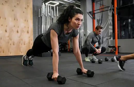 Woman doing dumbbell push-up in a gym with various equipment in the background.