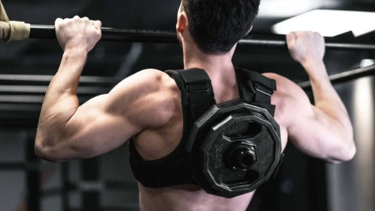 A person does a pull-up in a gym while wearing a weighted vest with a large weight plate attached to the back.